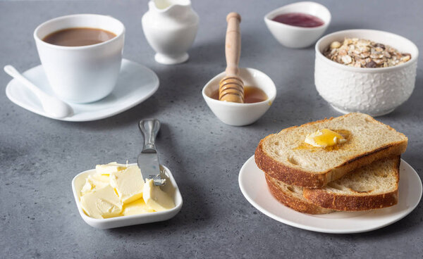 Butter and bread for breakfast with cup of coffee, granola, honey and jam over grey stone background. 