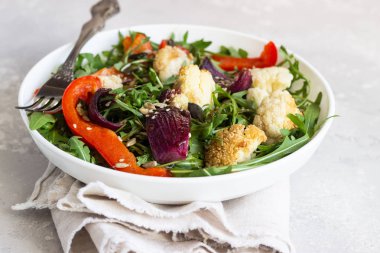 Roka, kavrulmuş sebze (karnabahar, kırmızı biber ve soğan) ve tohum salatası. Sağlıklı gıda. Açık gri beton renkli. Kopya alanı.