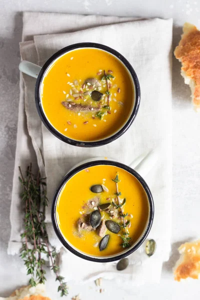 Two portions of fresh homemade pumpkin creamy soup served with liver and seeds in a white enamel mugs on a light grey concrete background. View from above, flat lay. — Stock Image Two Portions Fresh Homemade Pumpkin Creamy Soup Served Liver Seeds Royalty Free Stock Images