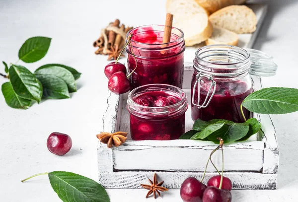 Homemade black cherry jam with spices (cinnamon, anise) with fresh cherries in the jars and fresh bread on a wooden cutting board. Light stone background. Homemade preserves.