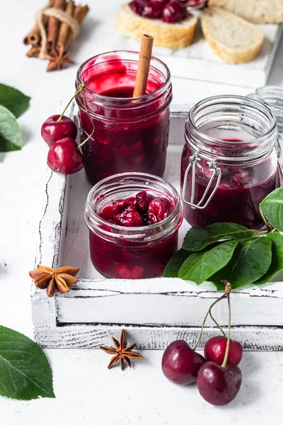 Homemade black cherry jam with spices (cinnamon, anise) with fresh cherries in the jars on a light stone background. Homemade preserves.