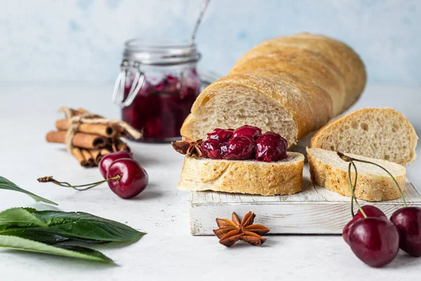 Homemade black cherry jam with spices (cinnamon, anise) with fresh cherries in the jars and fresh bread on a wooden cutting board. Light stone background. Homemade preserves.