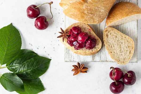 Homemade black cherry jam with spices (cinnamon, anise) with fresh cherries in the jars and fresh bread on a wooden cutting board. Light stone background. Homemade preserves.