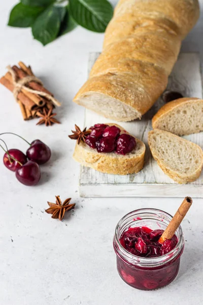 Homemade black cherry jam with spices (cinnamon, anise) with fresh cherries in the jars and fresh bread on a wooden cutting board. Light stone background. Homemade preserves.