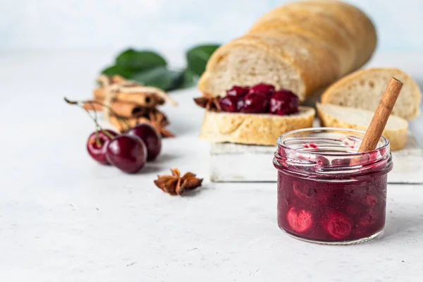 Homemade black cherry jam with spices (cinnamon, anise) with fresh cherries in the jars and fresh bread on a wooden cutting board. Light stone background. Homemade preserves.