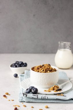 Kuru üzüm, taze yaban mersini ve süt ile beyaz bir seramik kase ev yapımı granola. Açık gri arka planda sağlıklı kahvaltı malzemeleri.