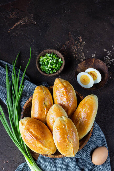 Traditional Russian pies with green onion and egg on wooden plate, dark rustic background. Russian pirozhki, homemade baked patties.