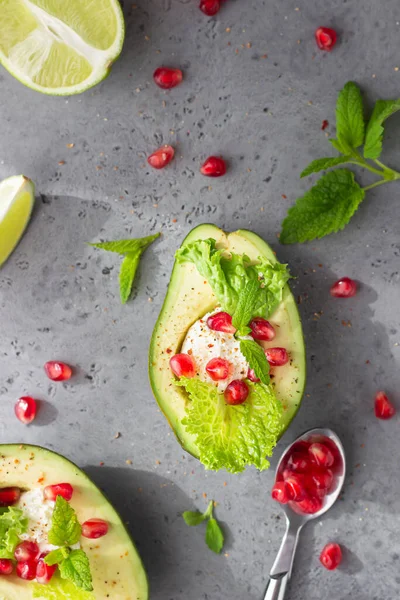 Halved avocado with cream cheese, pomegranate seeds, lettuce, mint and lime on grey stone background. Healthy food concept.