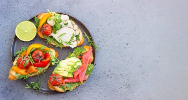 Izgara biber, domates, füme somon, hindi, avokado ve roka ile çeşitli kruvasan sandviçleri gri beton arka planda mikro yeşille servis edilir..