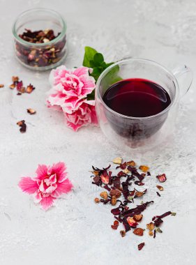 Cam bardakta sıcak amber çayı, pembe çiçekler ve açık gri arkaplanda kuru çay yaprakları..