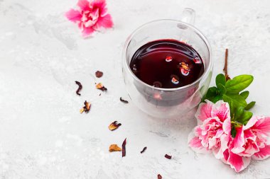 Cam bardakta sıcak amber çayı, pembe çiçekler ve açık gri arkaplanda kuru çay yaprakları..