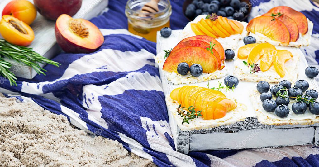 Healthy picnic for a summer vacation on the sea beach with fresh fruits and berries, sandwiches with cream cheese, fruits and blueberries with honey on a blue and white cloth.