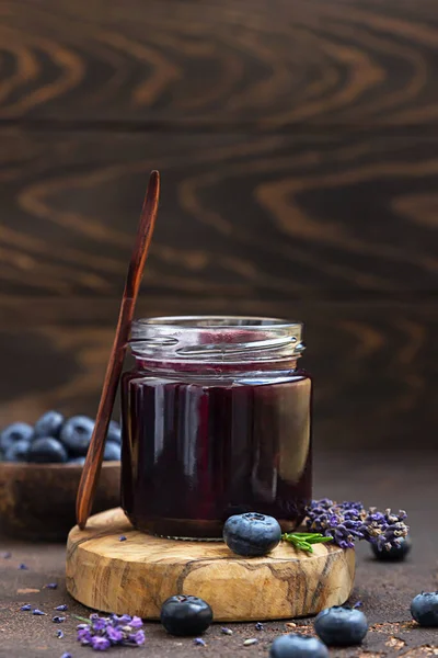 Glasburk och sked med läcker blåbärssylt med färska bär, lavendel och rosmarin på brunt bord. Hälsosam mat till frukost. — Stockfoto