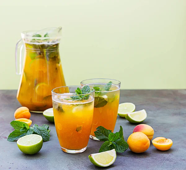 Delicious refreshing drink with apricot, lime and mint in jug and glasses on green background.