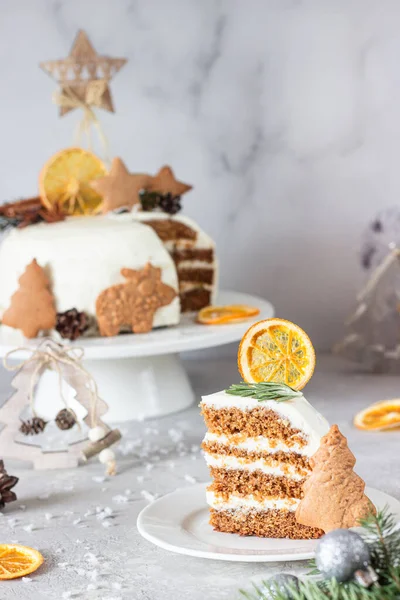 Christmas or New Year gingerbread cake with white cream decorated with gingerbread cookies, fir tree branches, cones, spices and dried orange slice. Winter cake. Grey concrete background.