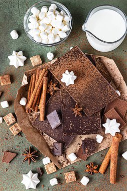 Sıcak çikolata yapmak için gerekli malzemeler. Koyu ve sütlü çikolata, kakao tozu, tarçın, anason yıldızı, esmer şeker, marşmelov, krema ve süt yeşil taş zemin üzerinde.