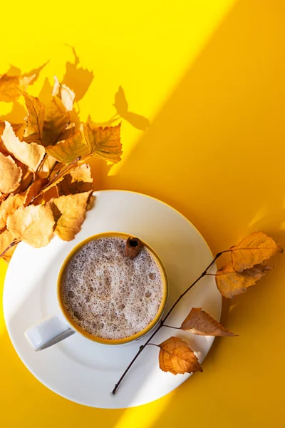 Autumn coffee or cappuccino composition. White coffee cup with milk foam, cinnamon stick and autumn leaves on yellow background. Fall hot drinks, cafe and bar concept. Bright sunlight and shadows.