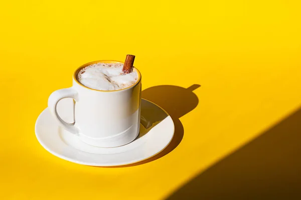 Weiße Keramiktasse Kaffee oder Cappuccino mit Zimtstange auf gelbem Hintergrund. Helles Sonnenlicht und harte Schatten. — Stockfoto