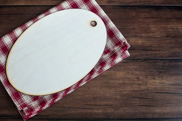 Empty wooden cutting board on a checkered red napkin on an old wooden brown background, top view. Image with copy space. Kitchen table with a towel and a board - top view with copy space.