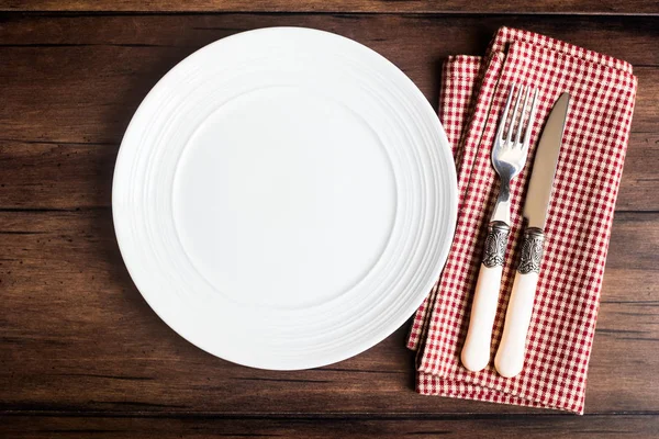Empty white plate, fork and knife on a checkered red napkin on an old wooden brown background, top view. Image with copy space. Kitchen table with a towel and a plate - top view with copy space.