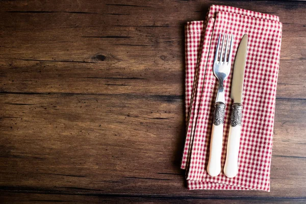 Fork and knife on a checkered red napkin on an old wooden brown background, top view. Image with copy space. Kitchen table with a towel and silverware - top view with copy space.