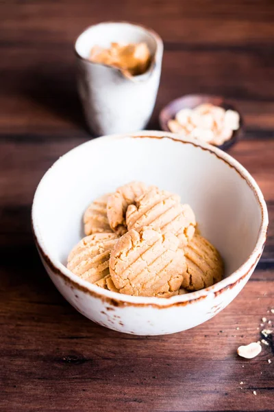 Heap of homemade peanut butter cookies in a bowl with a plate of peanuts and a cup of peanut butter on a wooden table, selective focus. Image with copy space. Traditional american treat.