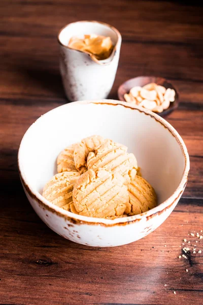 Heap of homemade peanut butter cookies in a bowl with a plate of peanuts and a cup of peanut butter on a wooden table, selective focus. Image with copy space. Traditional american treat.