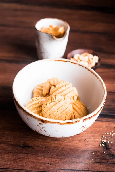 Heap of homemade peanut butter cookies in a bowl with a plate of peanuts and a cup of peanut butter on a wooden table, selective focus. Image with copy space. Traditional american treat.