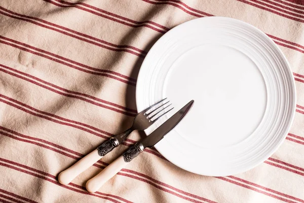 Empty white plate, fork and knife on a striped red napkin, top view. Image with copy space. Kitchen table with a towel and a plate - top view with copy space.