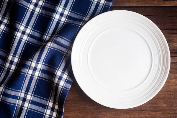 Empty white plate on a napkin on an old wooden brown background, top view. Image with copy space. Kitchen table with a towel and a plate - top view with copy space.