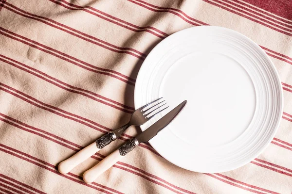 Empty white plate, fork and knife on a striped red napkin, top view. Image with copy space. Kitchen table with a towel and a plate - top view with copy space.