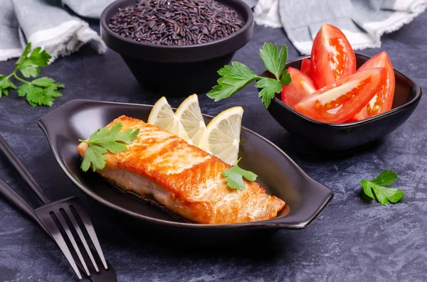 Roast salmon steak with unpolished rice and vegetables in a dish on a slate background. Selective focus.