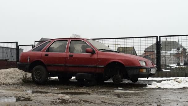 Een oude rode auto zonder wielen staat op de werf onder de eerste sneeuw — Stockvideo