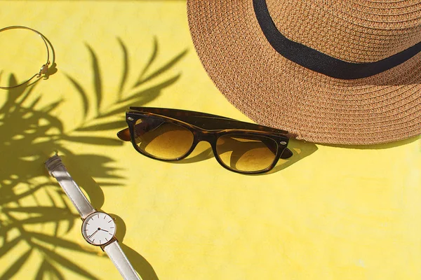 Female accessories Watch Sunglasses Straw hat Bracelet Summer holiday concept Flat lay Top view Yellow background Copy space Tropical leaf shadow Beautiful composition