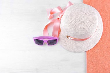Pembe kurdele ve turuncu bir havlu üzerinde pembe güneş gözlüğü ile bir yaz şapka. Beyaz ahşap arka plan, üstten görünüm.