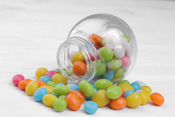 Multicolored round candy in a glass jar on a white wooden background.