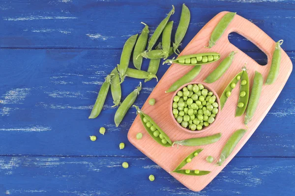 Fresh, green peas in a wooden bowl on a wooden board next to peas in a stitch on a blue wooden background. top view