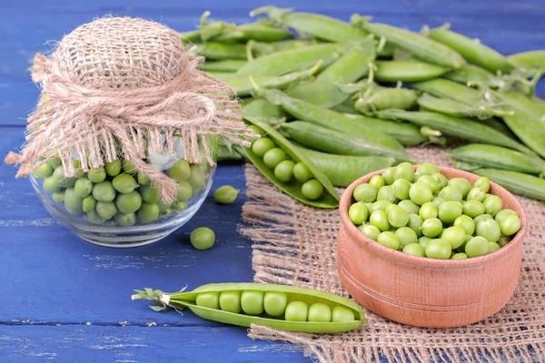 Fresh green peas in a wooden bowl next to peas in a stitch on a blue wooden background