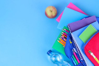 Çok çok renkli okul malzemeleri mavi okul sırt çantasından parlak mavi bir arka plan üzerinde düşüyor. Üstten görünüm