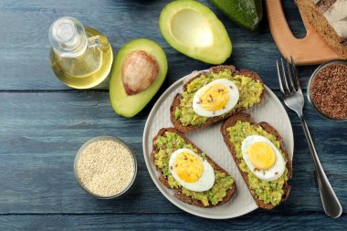 Avokado püresi ve bir tabak ve üzerinde mavi bir ahşap masa yemek pişirmek için malzemeler üzerinde yumurta ile sandviç. yukarıdan görüntülemek