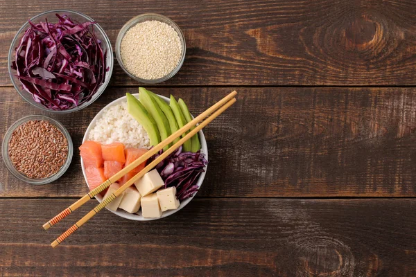 poke bowl Hawaiian food. a plate with rice, salmon, avocado, cabbage and cheese, near sesame. on a brown wooden table. view from above