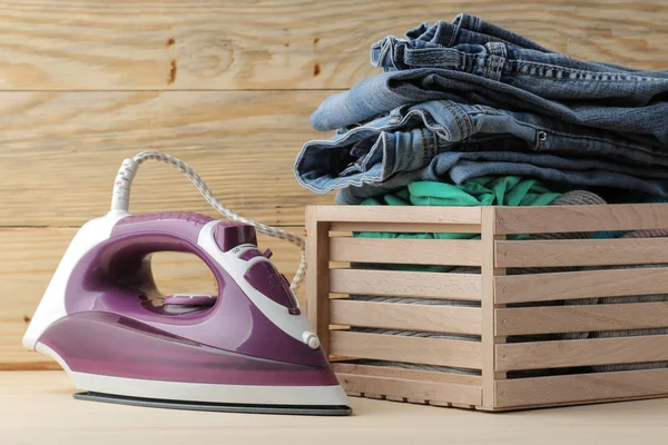 lilac iron and a stack of clothes in a box on a natural wooden background. ironing clothes. household electrical appliances.