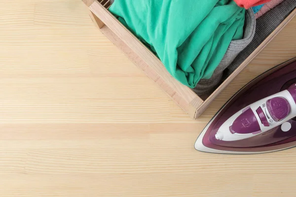 lilac iron and a stack of clothes in a box on a natural wooden background. top view with place for text. ironing clothes. household electrical appliances.