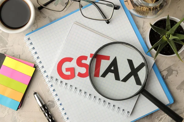 The word GST TAX with a notebook, glasses, pen and coins in a can on a light concrete background. view from above