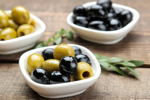 Green and black olives with leaves in a white bowl on a brown wooden table. close-up