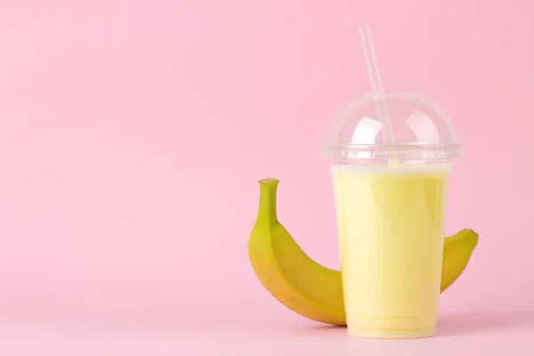 a plastic disposable glass with a banana milkshake and a fresh banana on a bright trendy pink background. Making a milkshake. space for text