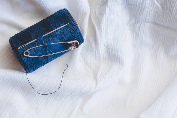 Blue thread spool with sewing needle and pin on white cotton cloth