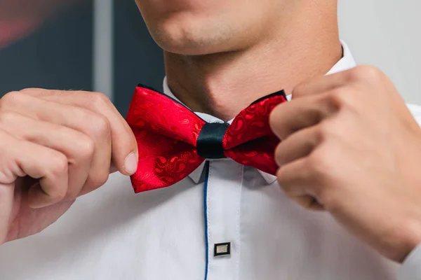 A man in a white shirt ties a red bow tie while preparing for a wedding ceremony close up
