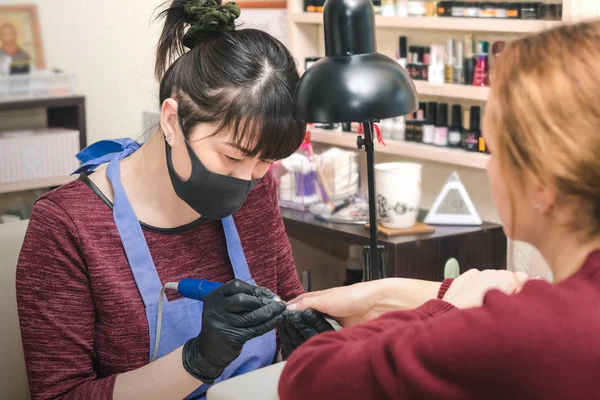 Güzellik salonunda bir el ile elektrikli freze makinesi kullanarak istemciye bir manikür manikürcü Siyah lateks eldiven yapar.