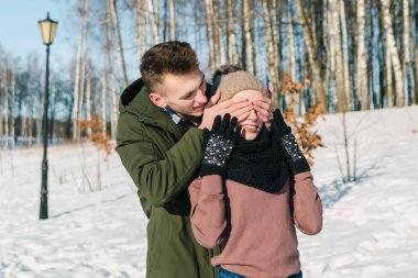 Güzel genç çift aşık bir açık güneşli kış gününde parkta yürüyor. Adam sevgilisinin gözleri elleriyle kapatır.